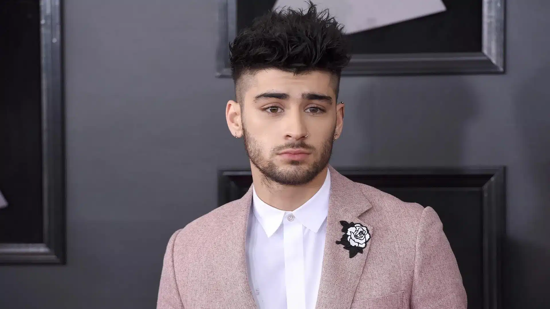 Zayn Malik poses wearing a dusty rose blazer with a black and white rose lapel pin and a crisp white button-down shirt against a dark gray backdrop