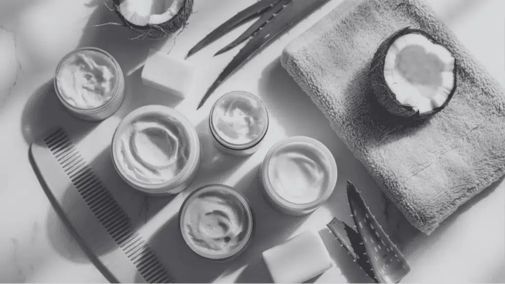 Flat lay of natural hair care cream jars, coconut, aloe, soap bars, wooden comb on white marble