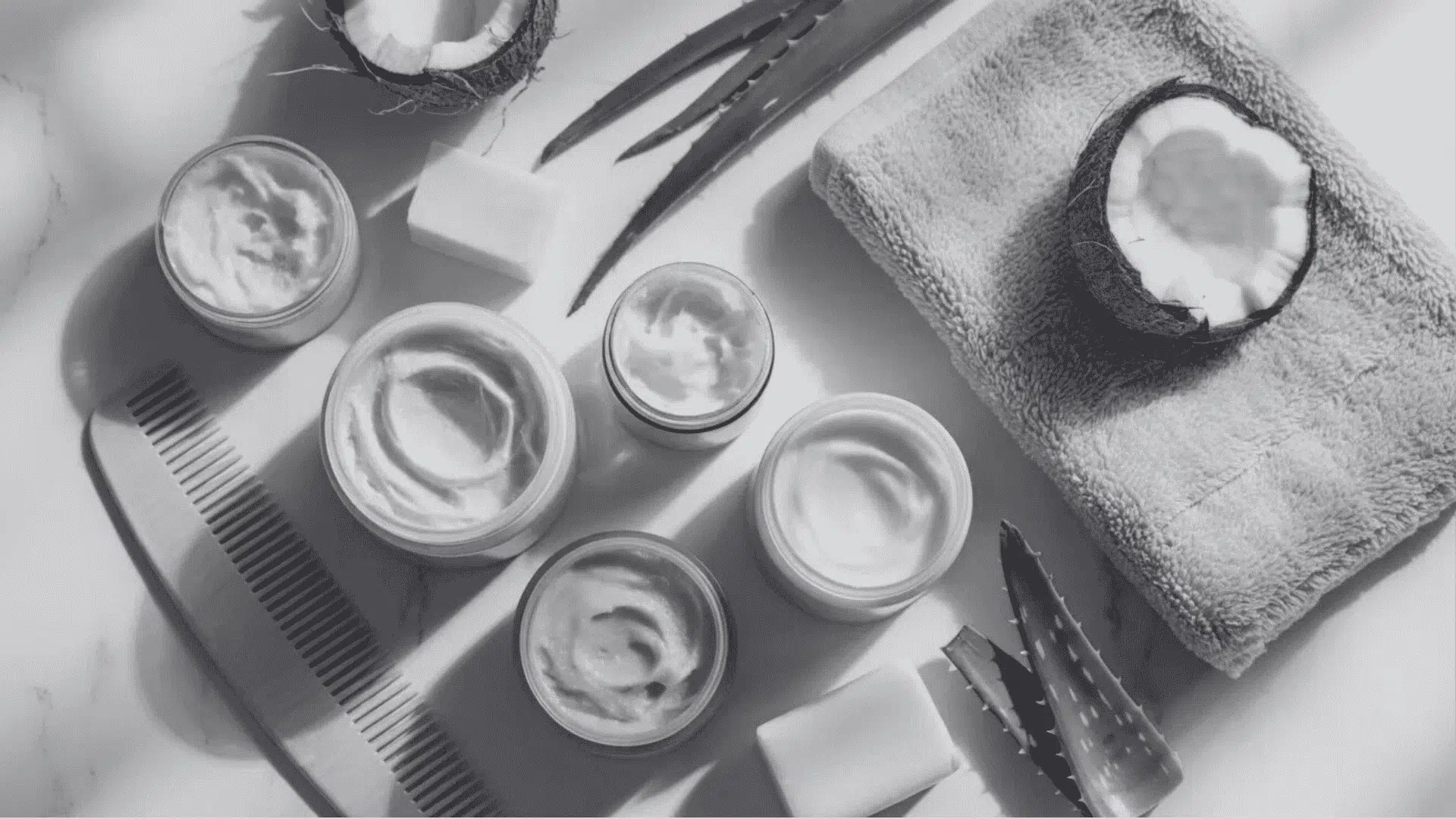 Flat lay of natural hair care cream jars, coconut, aloe, soap bars, wooden comb on white marble