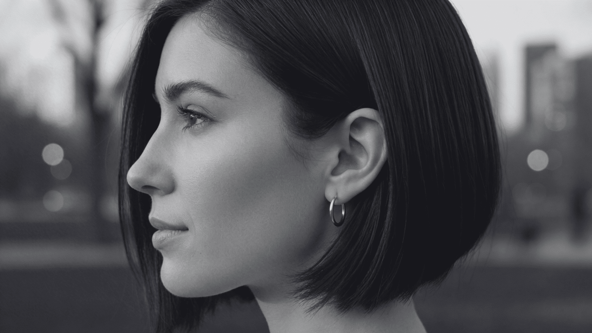 Profile view of a woman with short, dark brown bob haircut wearing silver hoop earrings outdoors in a park with blurred city skyline background