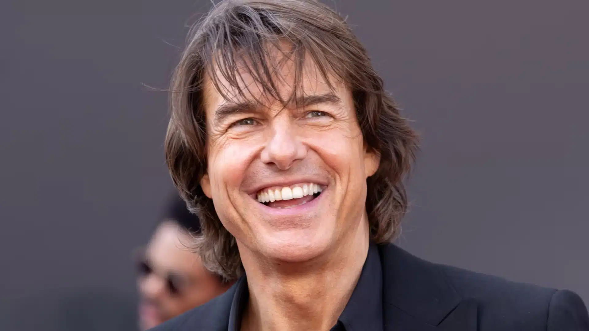 Tom Cruise smiles in a black suit, showcasing his long hair in a confident and stylish pose
