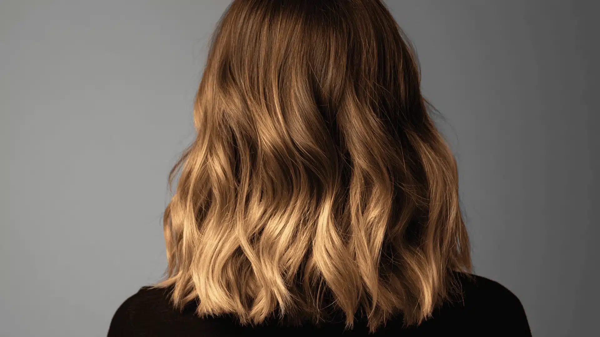 View from behind of a person with medium-length wavy hair featuring a warm brown to golden blonde with ombre gradient against a plain gray background