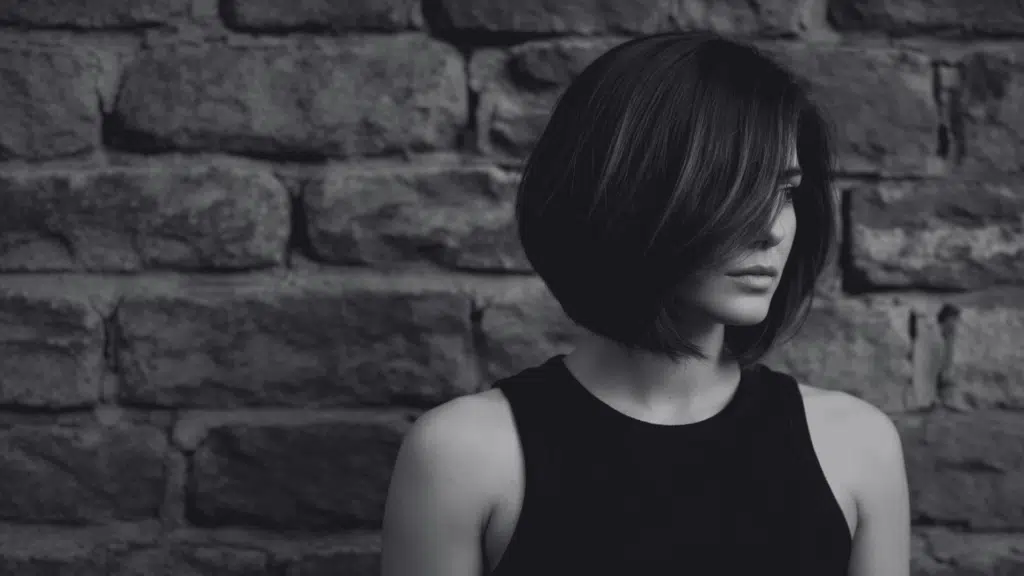 Woman with dark, layered, asymmetrical bob hairstyle wearing a black tank top against a rough-hewn stone wall background