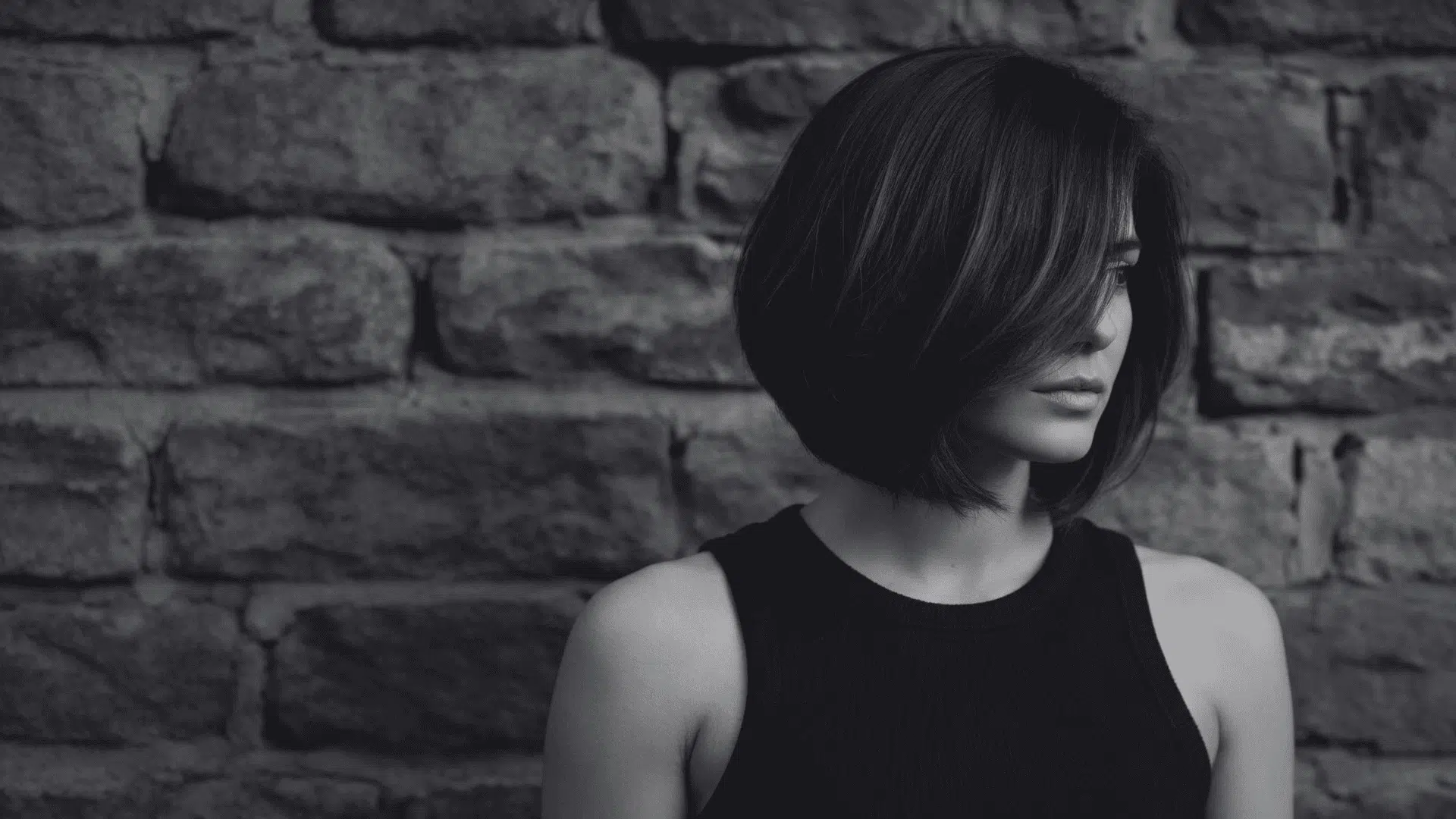 Woman with dark, layered, asymmetrical bob hairstyle wearing a black tank top against a rough-hewn stone wall background