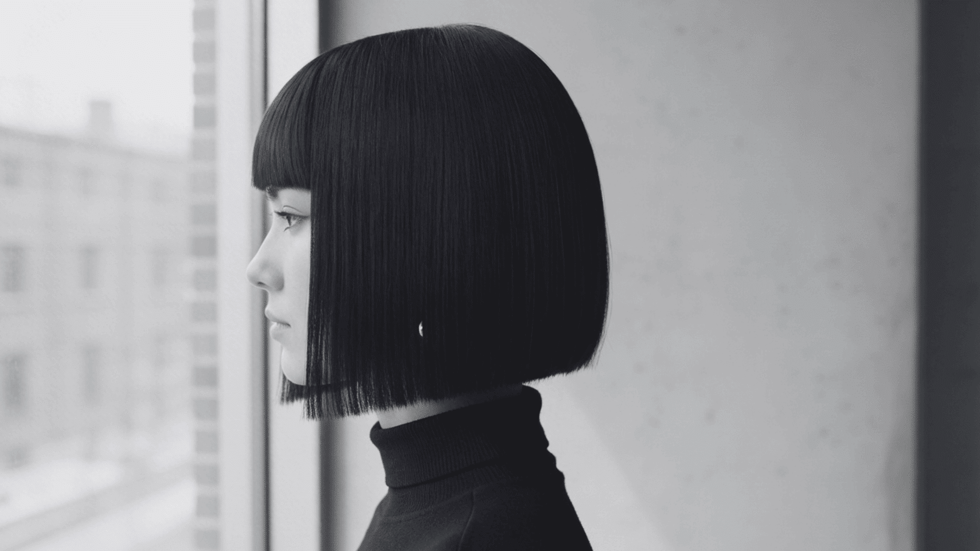 Woman with sharp black bob haircut and black turtleneck looking out a window at a blurred city scene