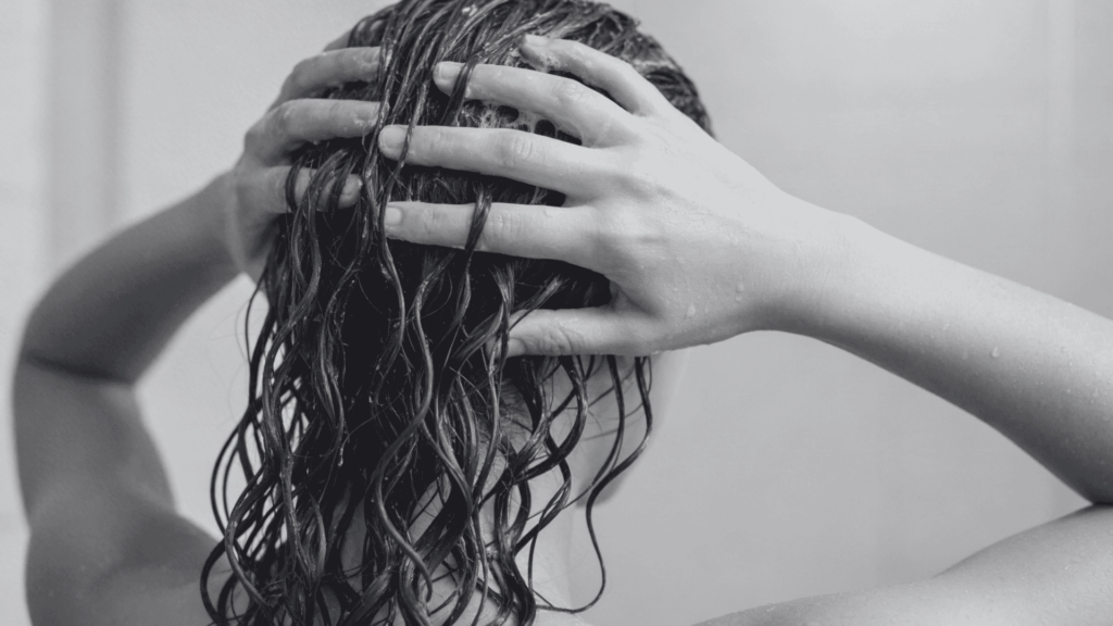 Woman with wet, dark, curly hair lathering conditioner with both hands during a shower