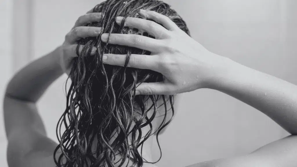 Woman with wet, dark, curly hair lathering conditioner with both hands during a shower