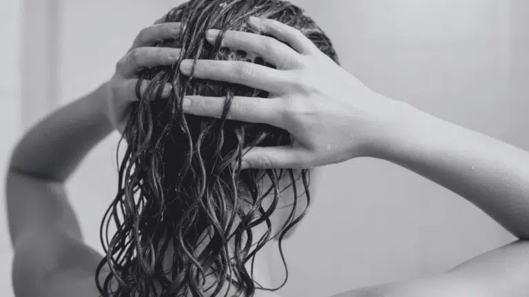 Woman with wet, dark, curly hair lathering conditioner with both hands during a shower