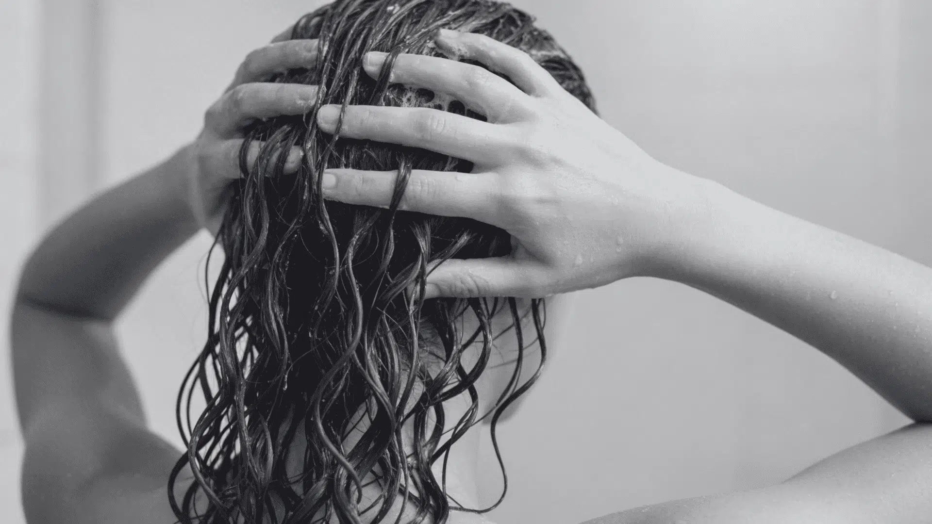 Woman with wet, dark, curly hair lathering conditioner with both hands during a shower