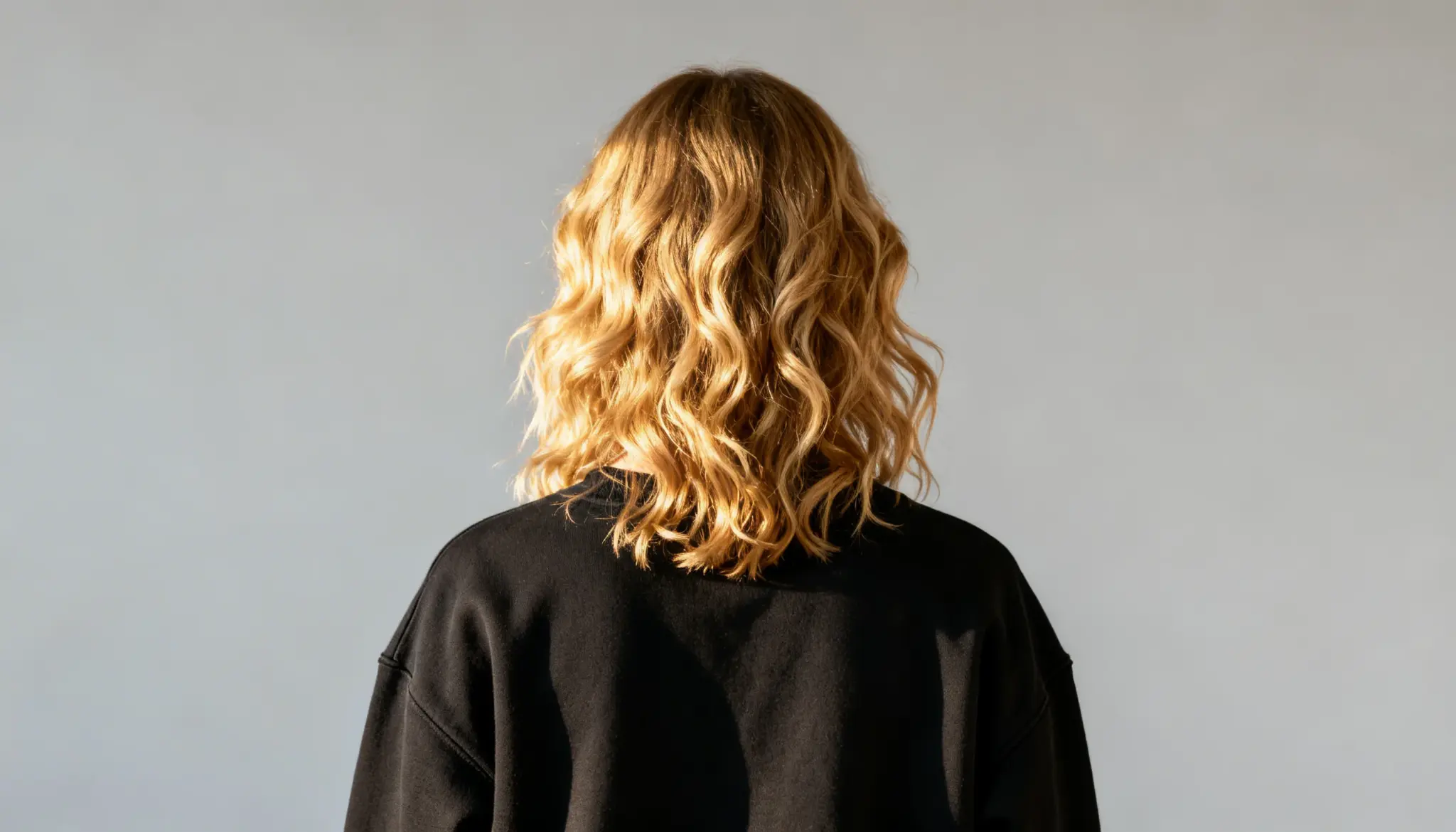 A person with wavy, blonde hair is seen from behind, wearing a black top against a plain gray background. The lighting highlights the hair’s texture.