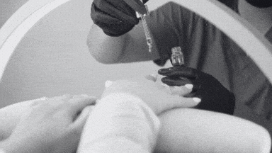 Black and white close-up of a gloved person applying cuticle oil from a dropper onto other person's nail