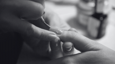 Close-up black and white shot showing a manicurist applying clear gloss to a client’s fingernail with a small brush