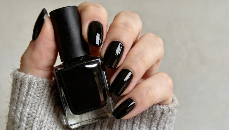 A hand with a freshly done gel manicure, holding a bottle of black gel nail paint while wearing a grey knit sweater cuff