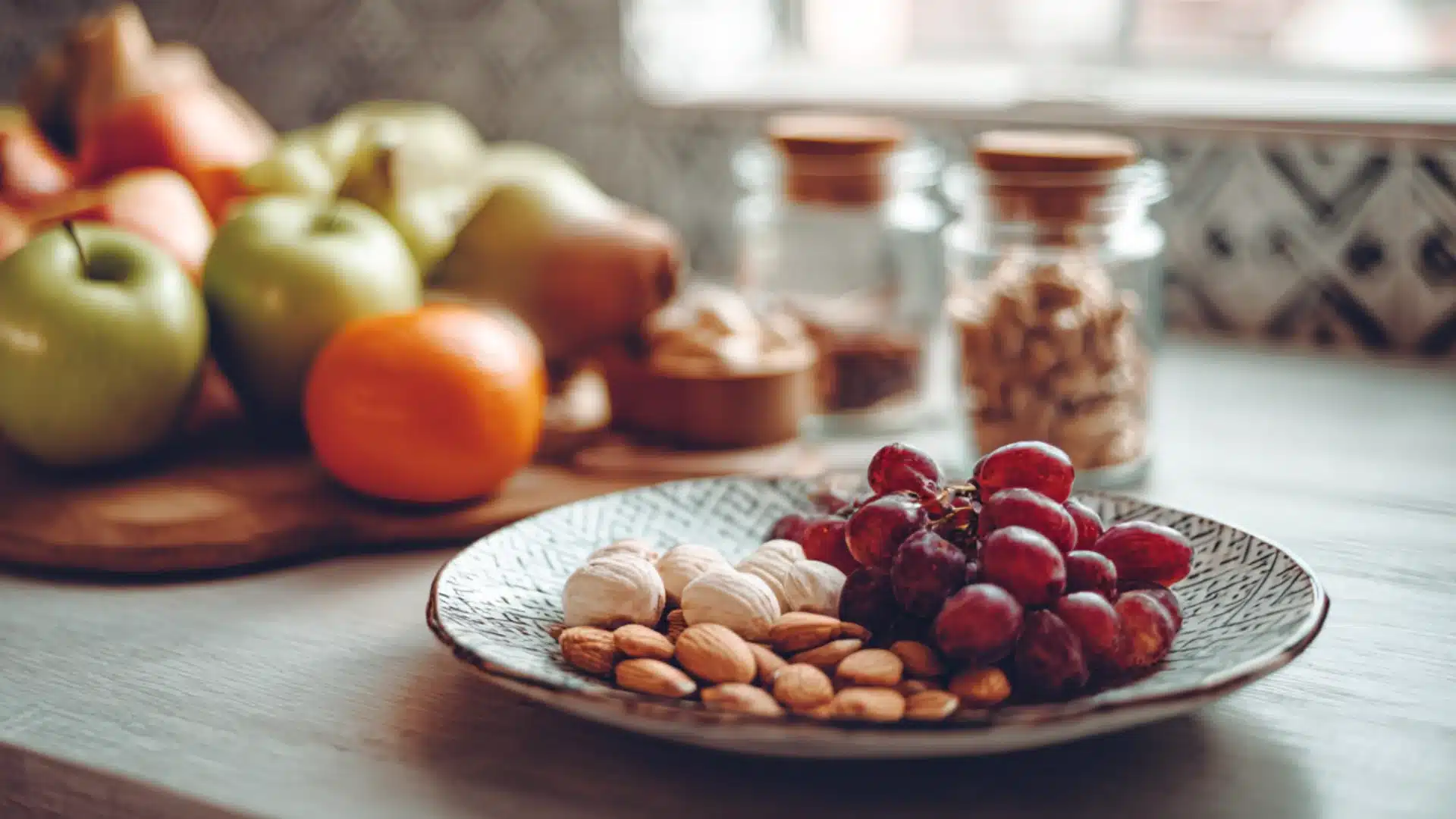 A plate of grapes and almonds sits on a kitchen counter near a bowl of apples and oranges and glass jars in the sunlight (1)