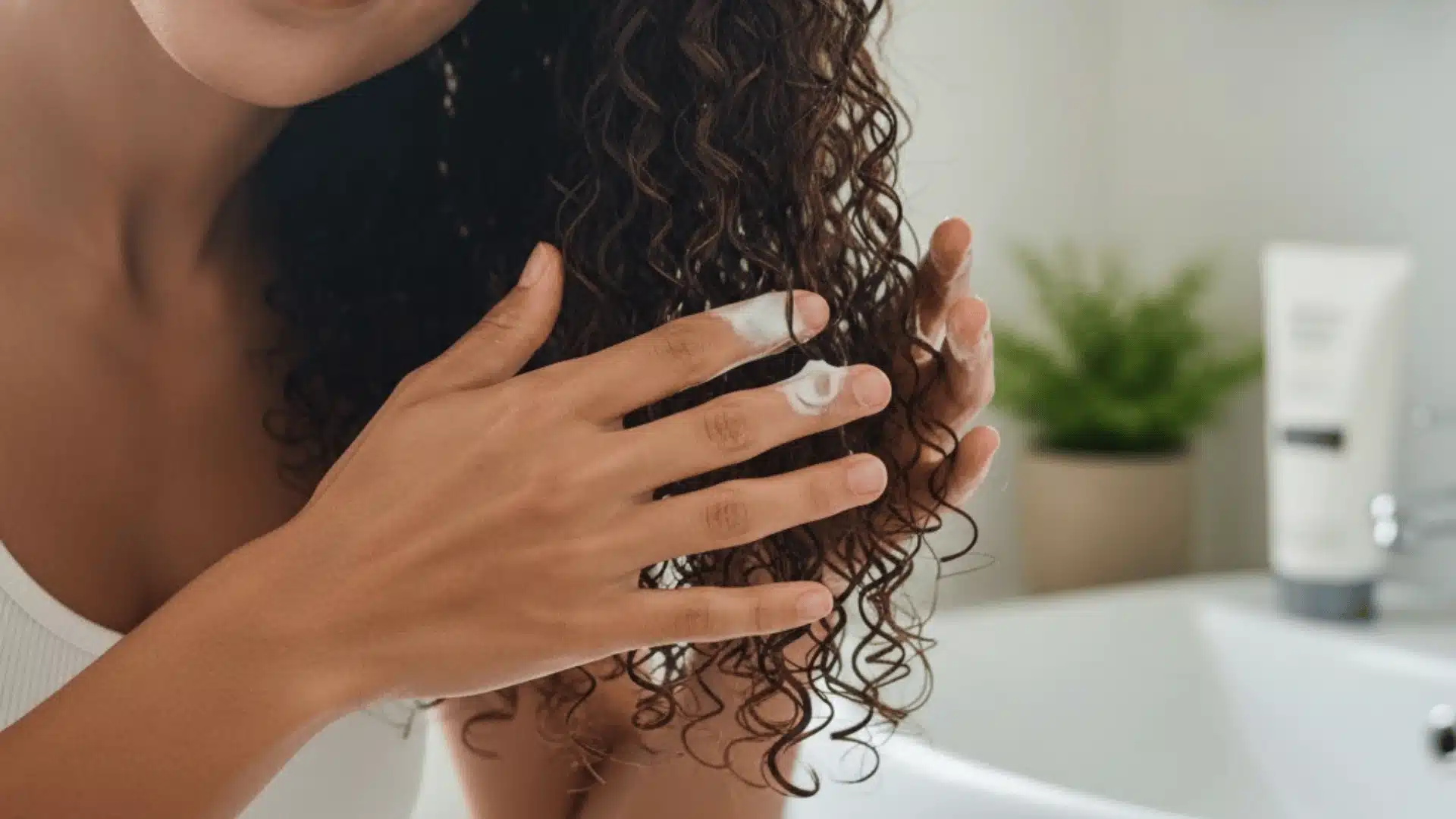 A woman applies conditioner to her wet, dark brown curly hair over a bathroom sink