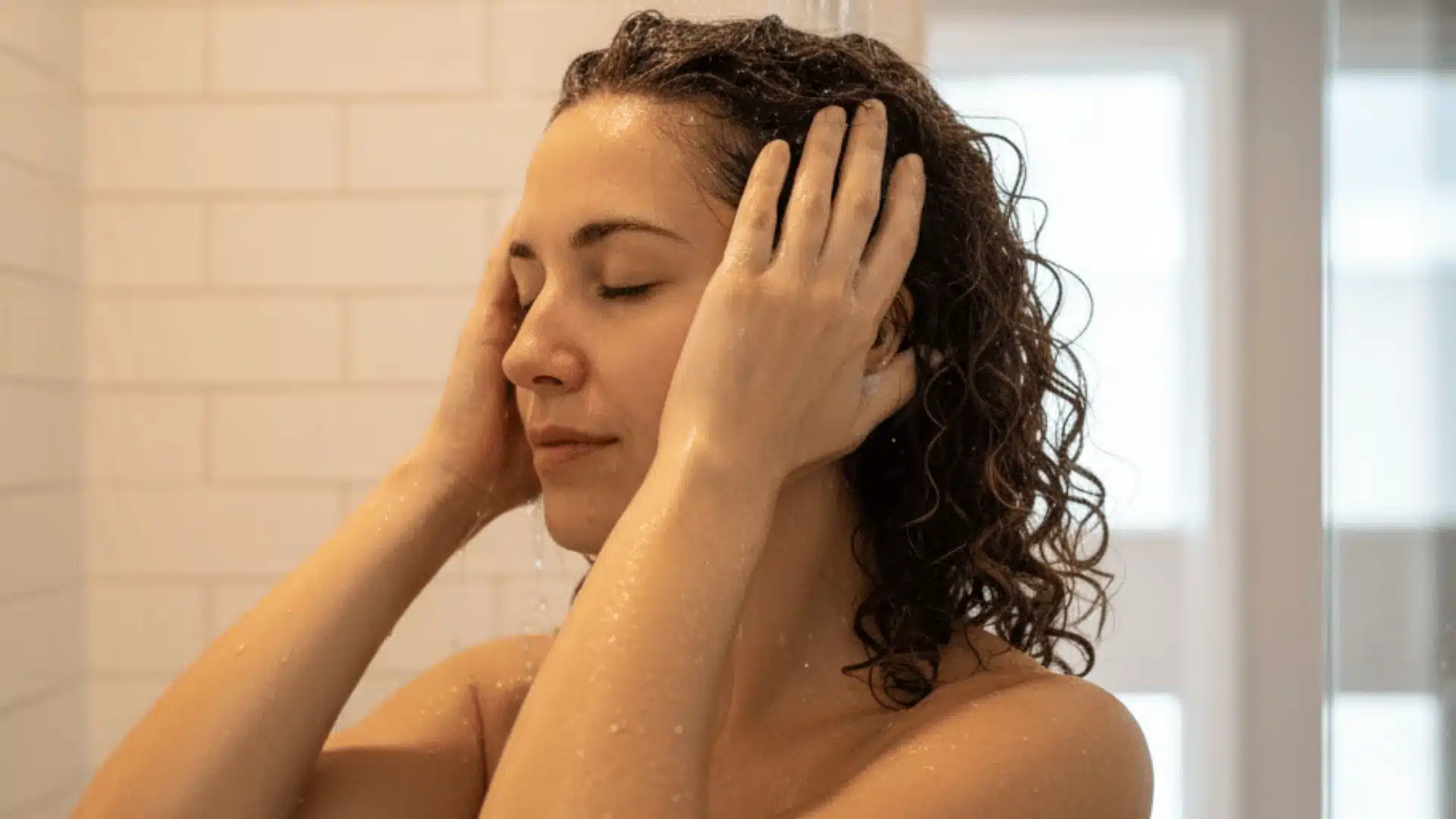 A woman with her eyes closed and wet curly brown hair while showering against a white subway tile background