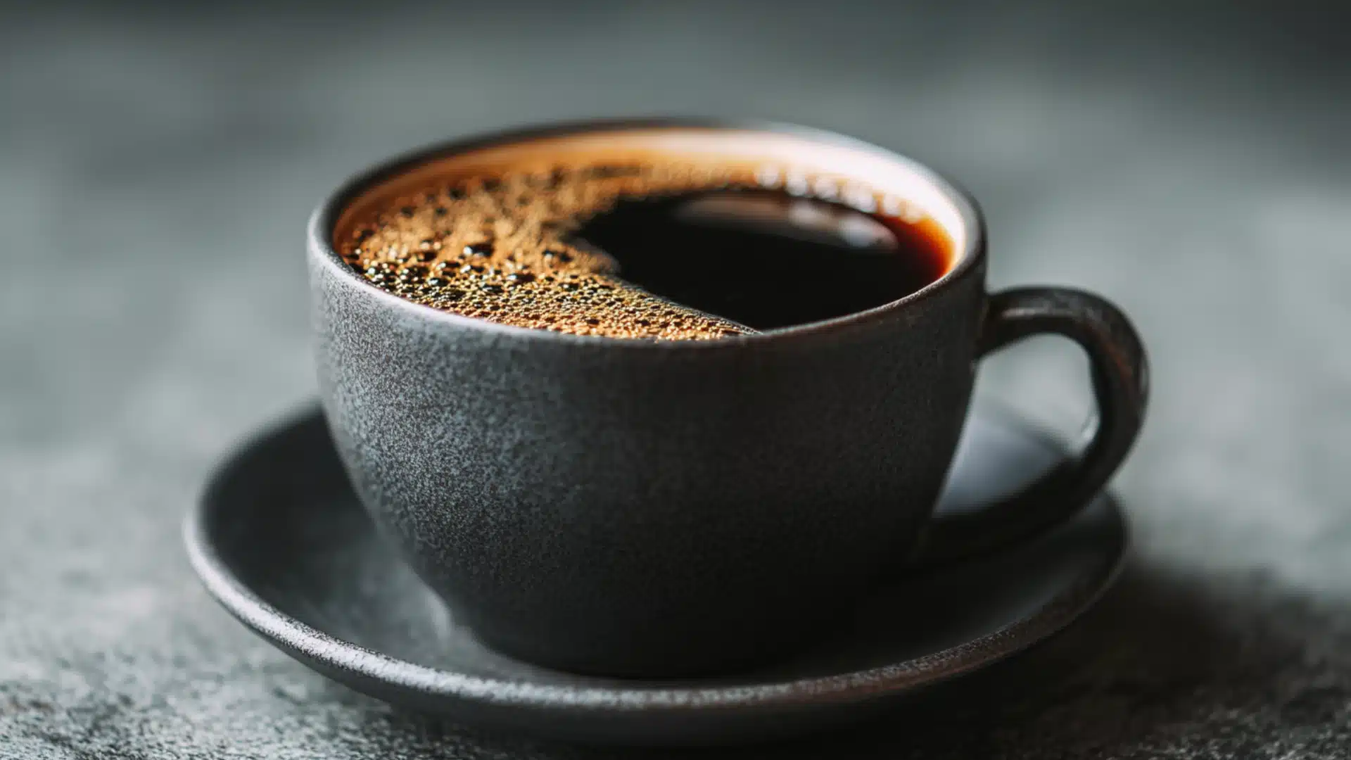 black textured mug filled with dark coffee and golden foam on a saucer calories in coffee shown in a close up side view