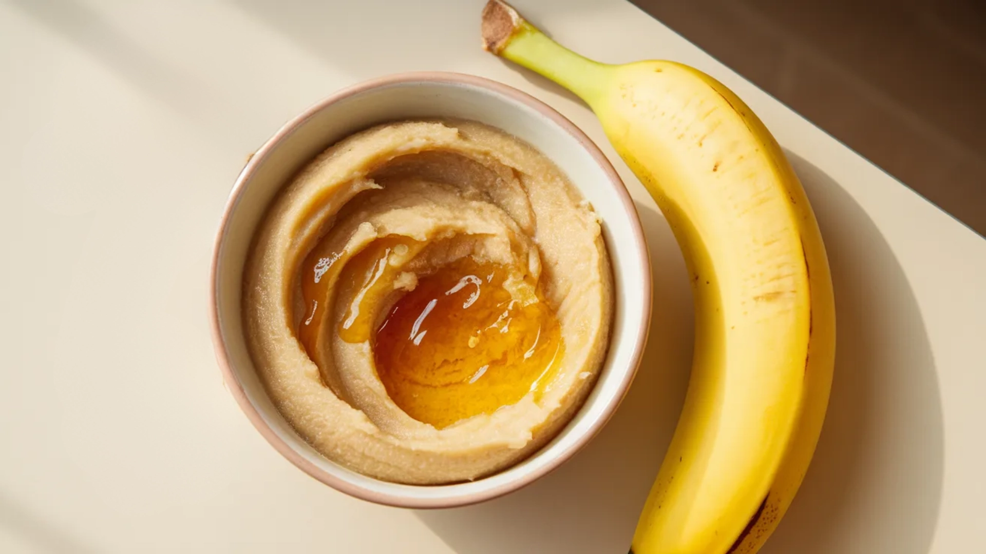 Mashed banana with honey in bowl, whole banana and coconut cream placed beside in sunlight