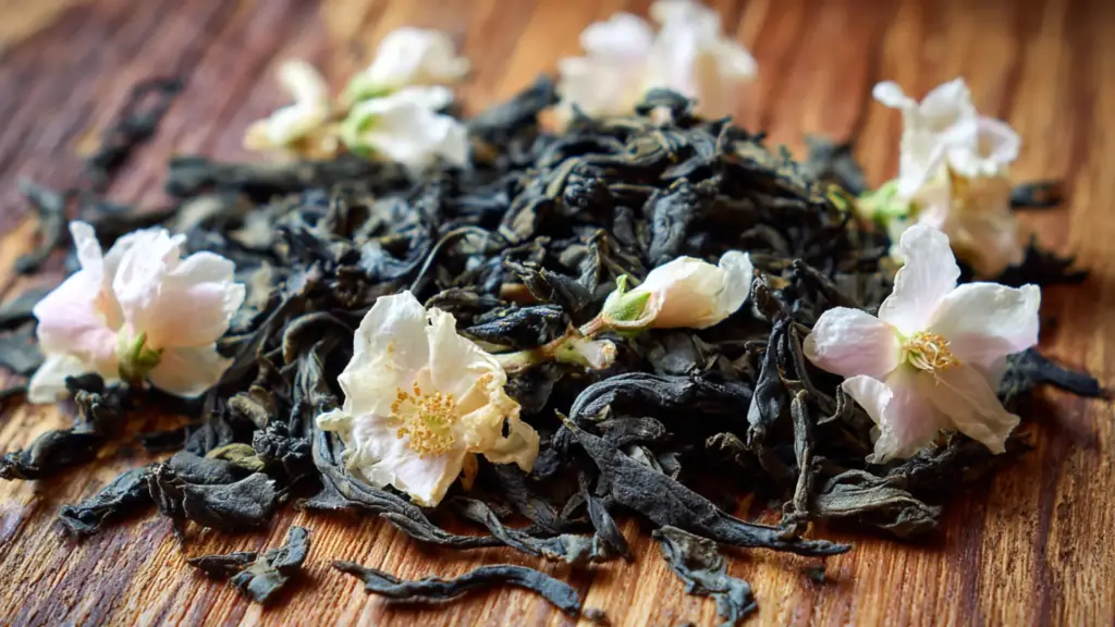 pile of dried green tea leaves mixed with small white jasmine flowers on a textured wood surface in a close up photo view