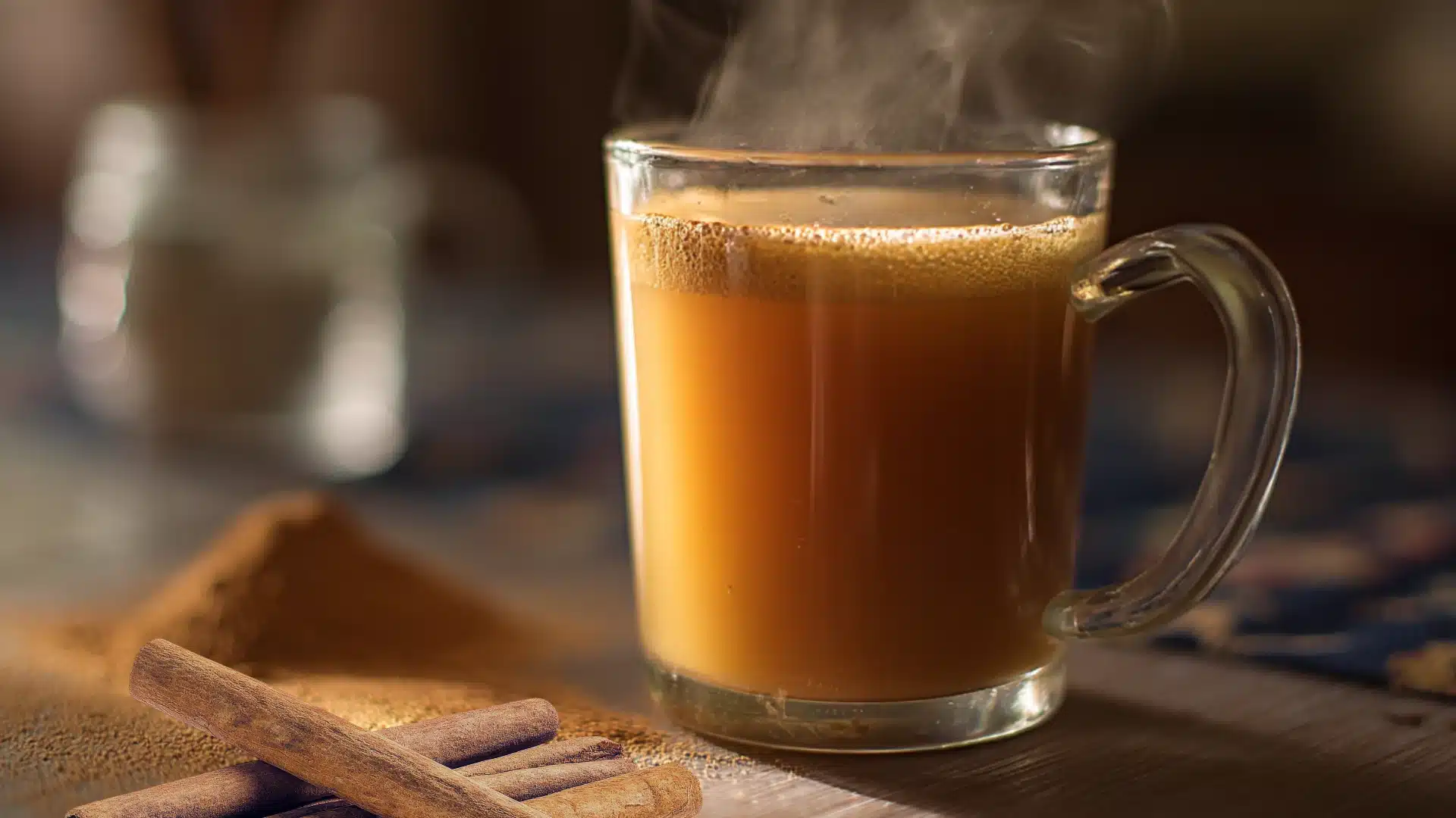 steaming glass mug of milky tea on a table with cinnamon powder in front and a blurry jar behind in warm morning light