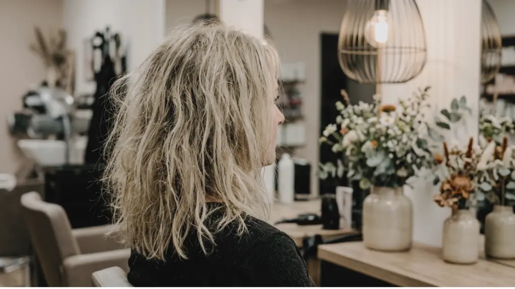 Woman with wavy blonde hair sitting in profile inside a softly lit salon with floral arrangements on a counter