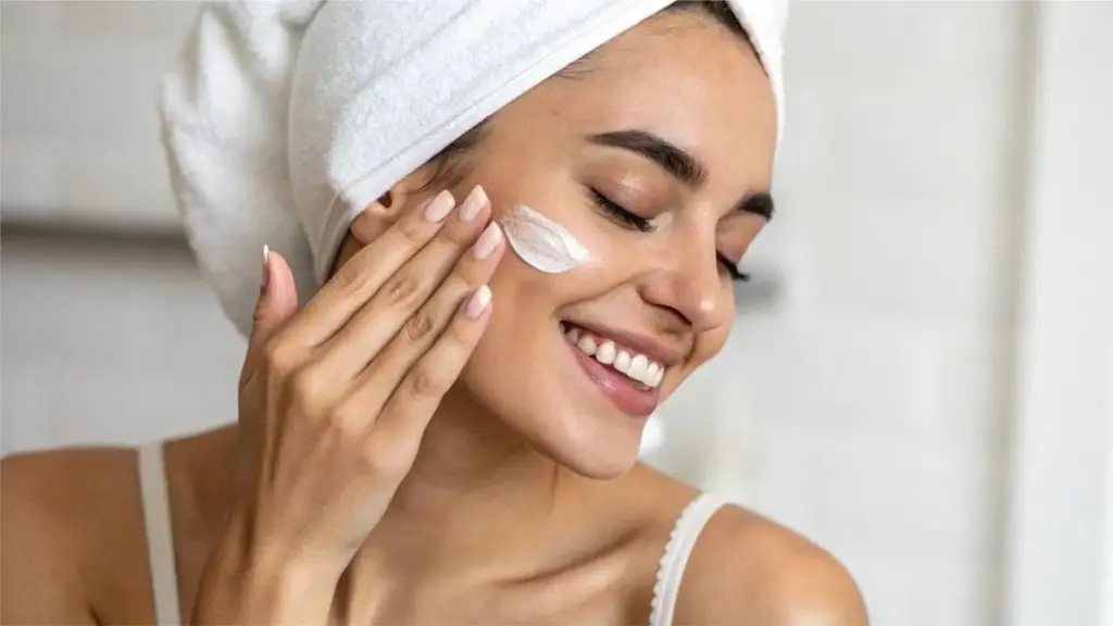 Woman applying facial cream in bathroom with white towel wrapped around hair