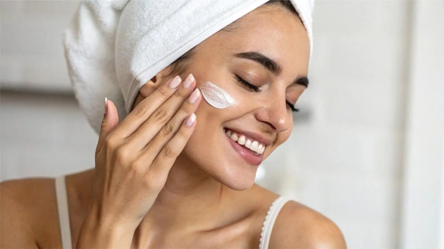 Woman applying facial cream in bathroom with white towel wrapped around hair