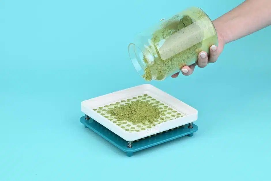 A hand pouring green herbal powder into a manual capsule filling tray