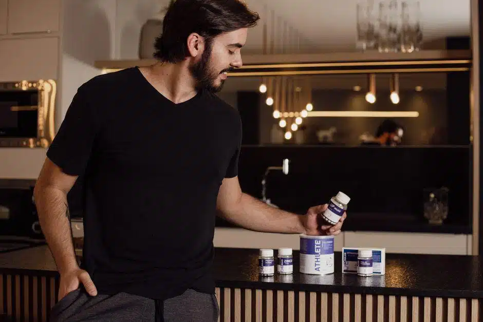 A man reading a supplement bottle label at a kitchen counter beside other bottles