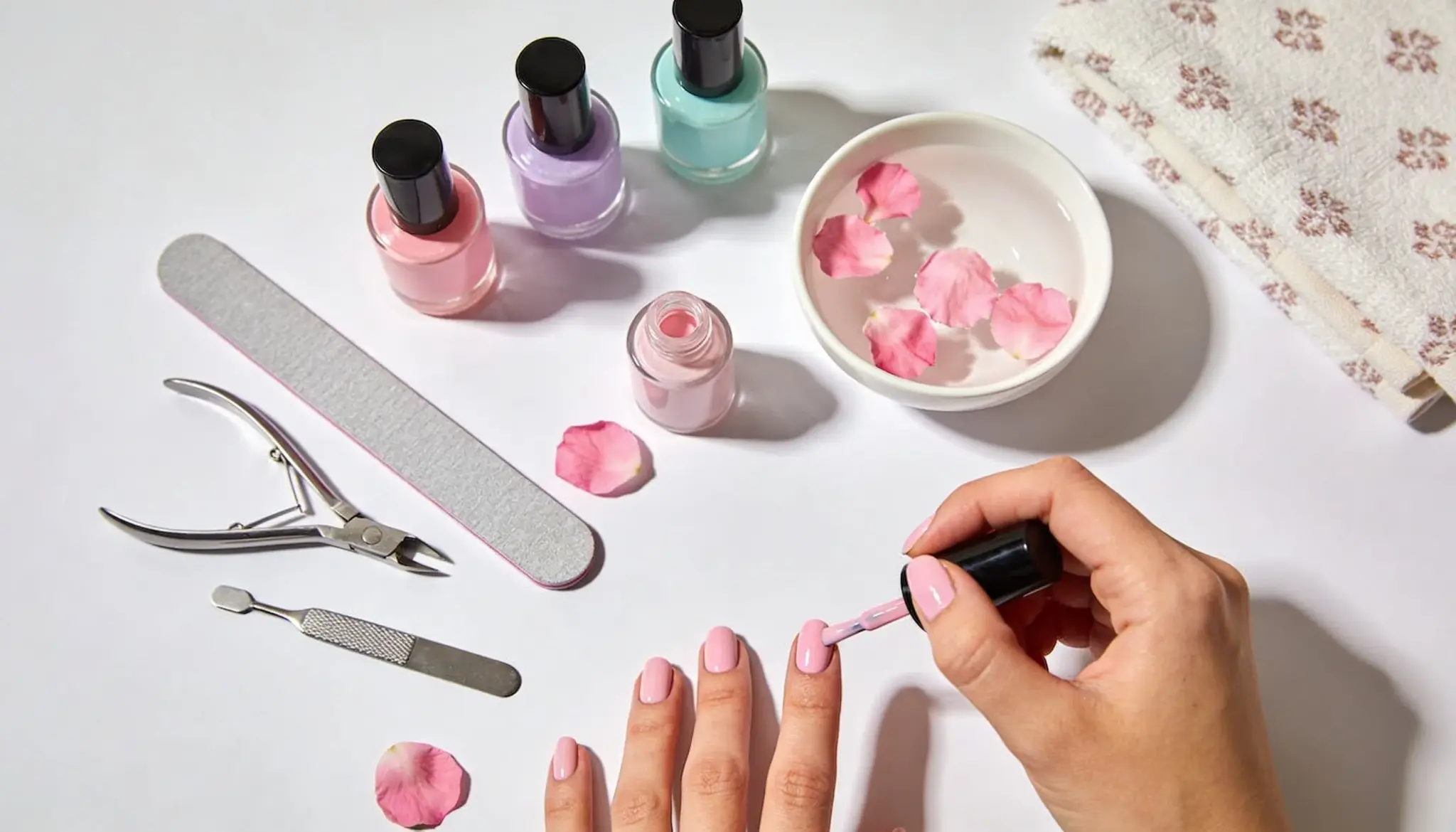 A person applies light pink nail polish while various manicure tools pastel polish bottles and a bowl of water with rose petals sit on a white surface