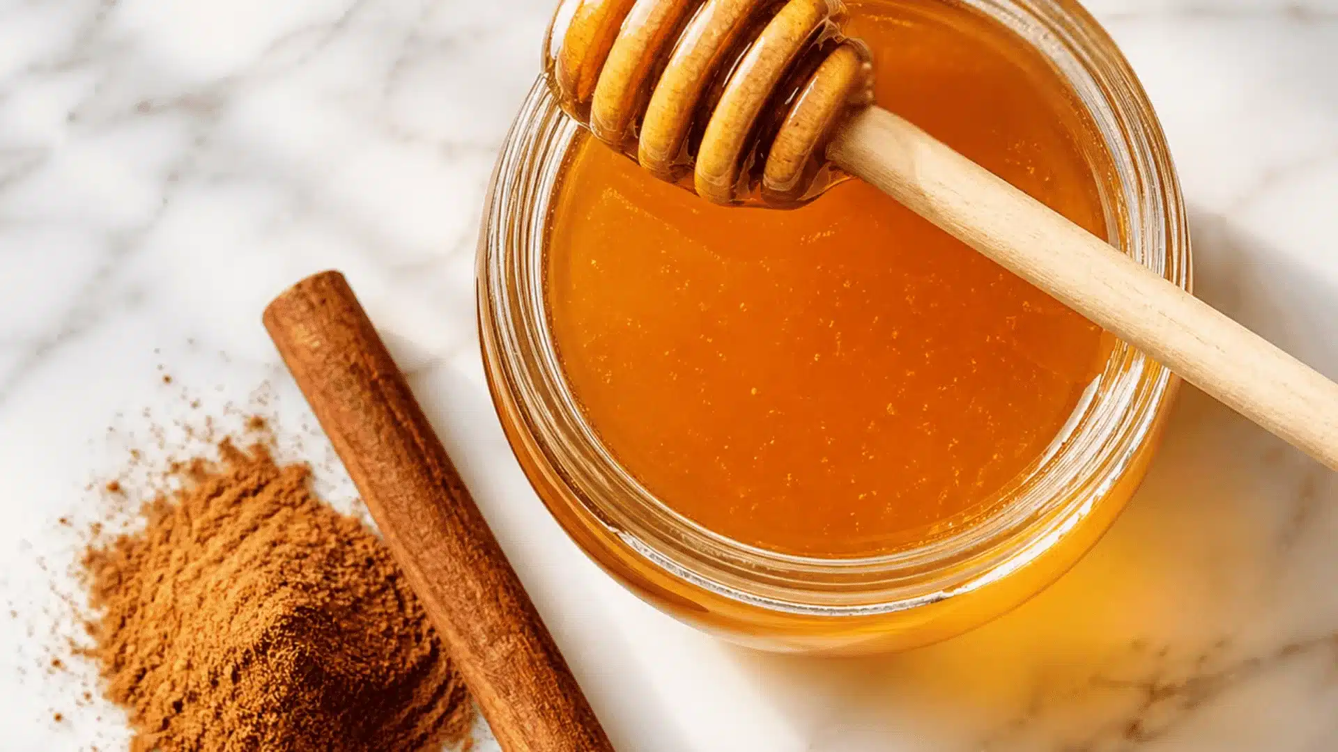 Glass jar of raw honey with dipper, cinnamon stick and powder on white marble, bright natural light.
