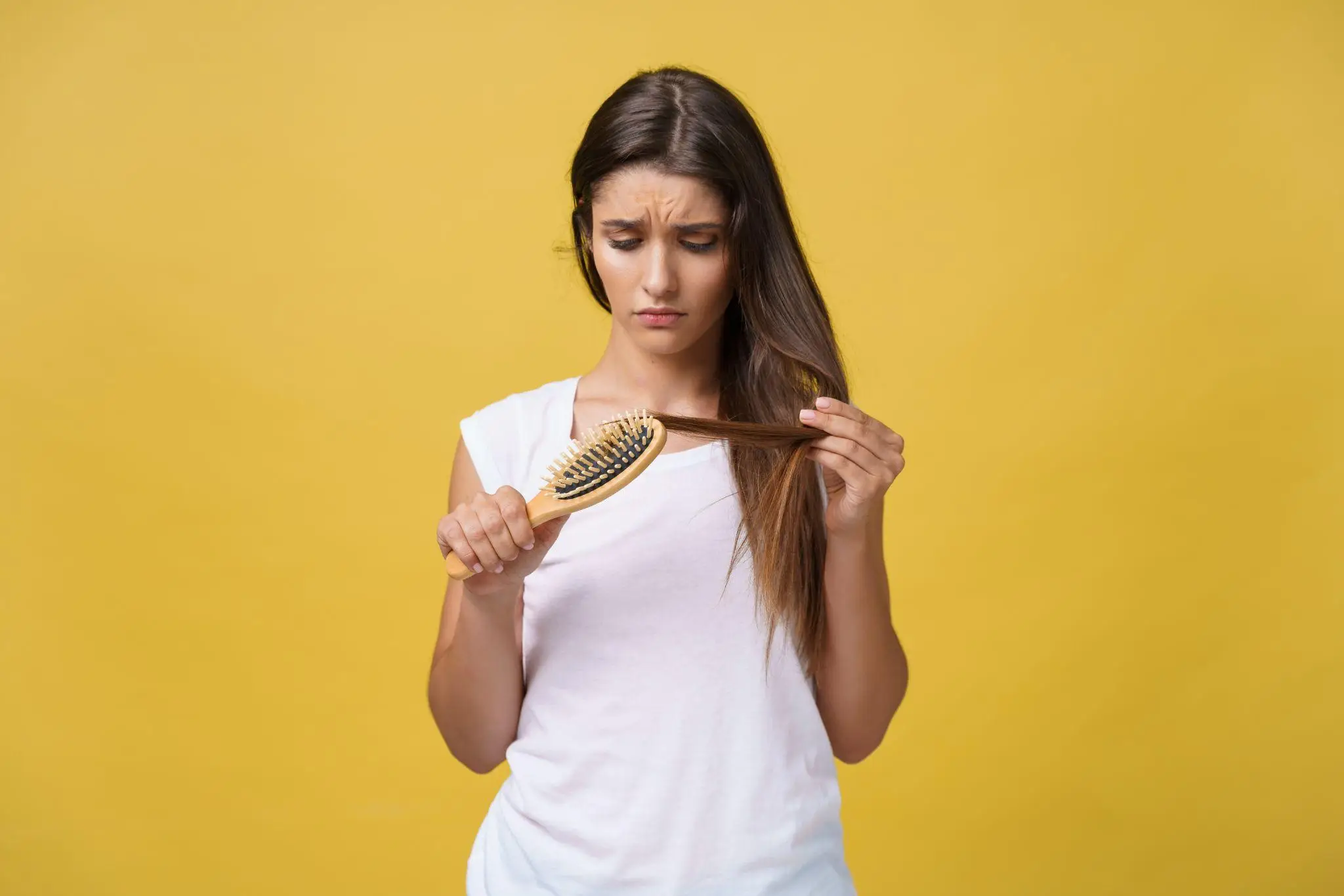 Woman examining hair and holding wooden brush against yellow background