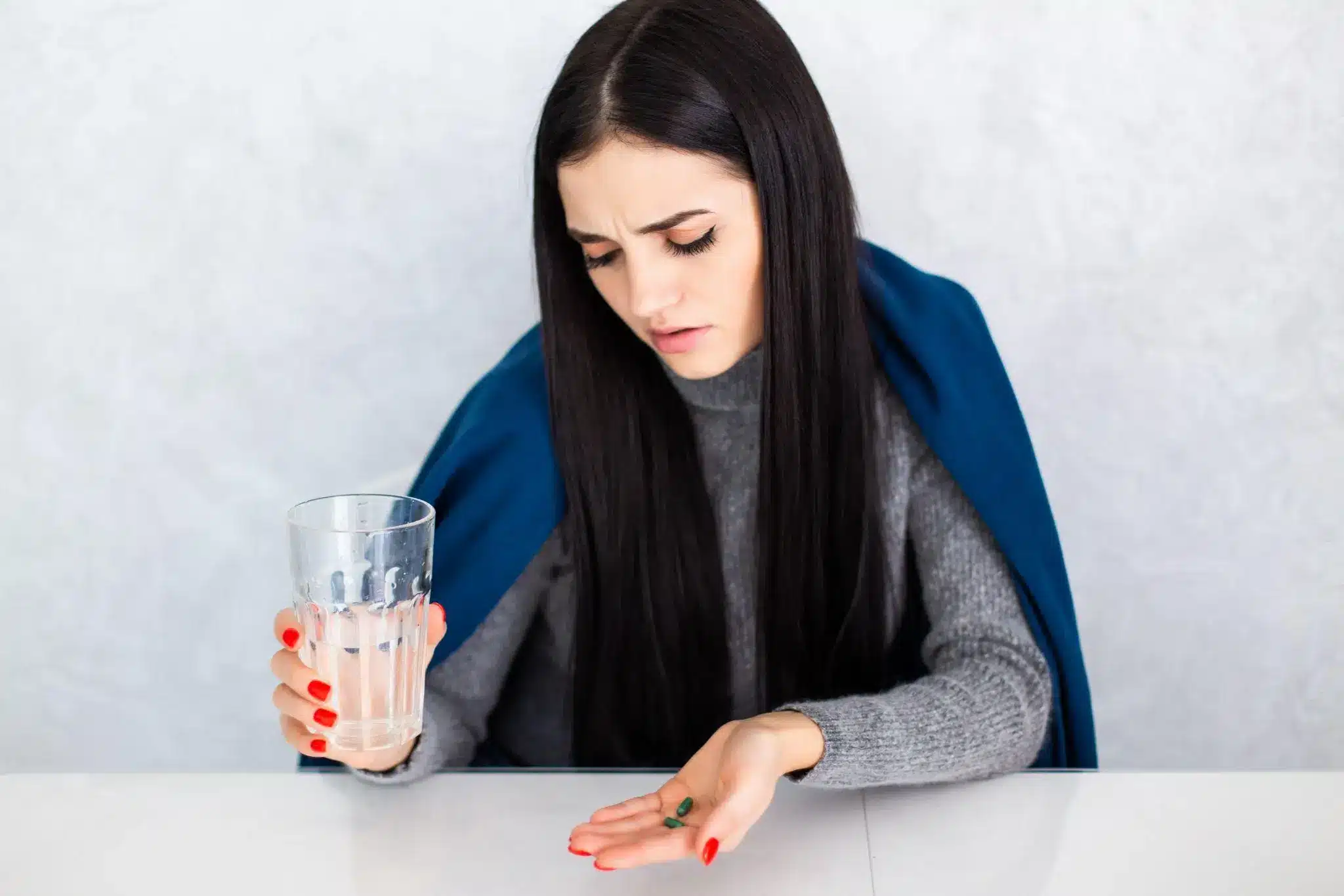 Woman in gray sweater holding glass of water and pills at white table