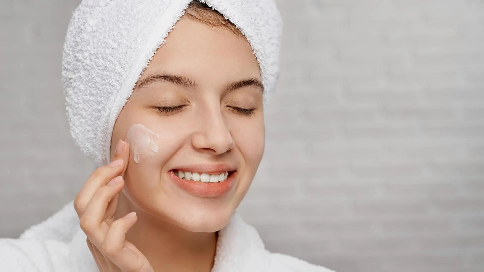 Young woman applying face cream with towel wrap in bathroom setting