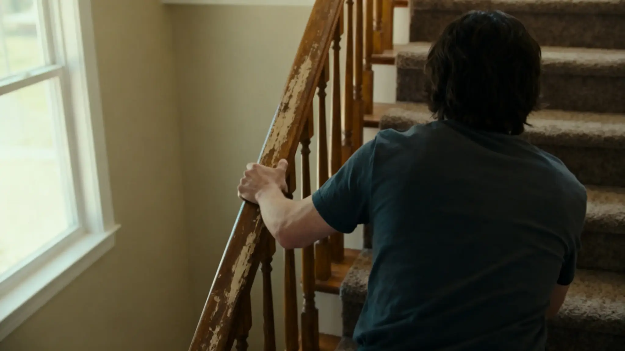 Person climbing carpeted stairs with hand on worn wooden railing in indoor setting
