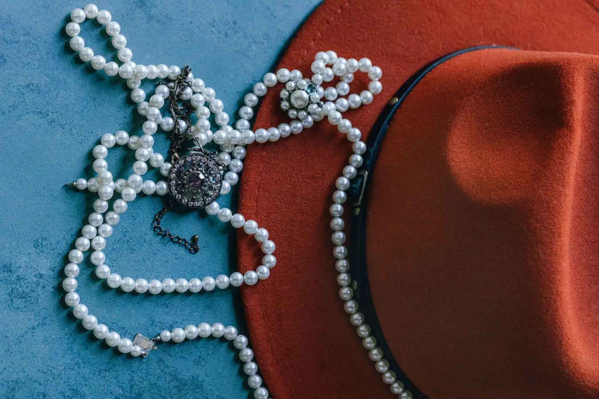 Pearl necklaces and ornate pendant on red felt hat against blue textured background