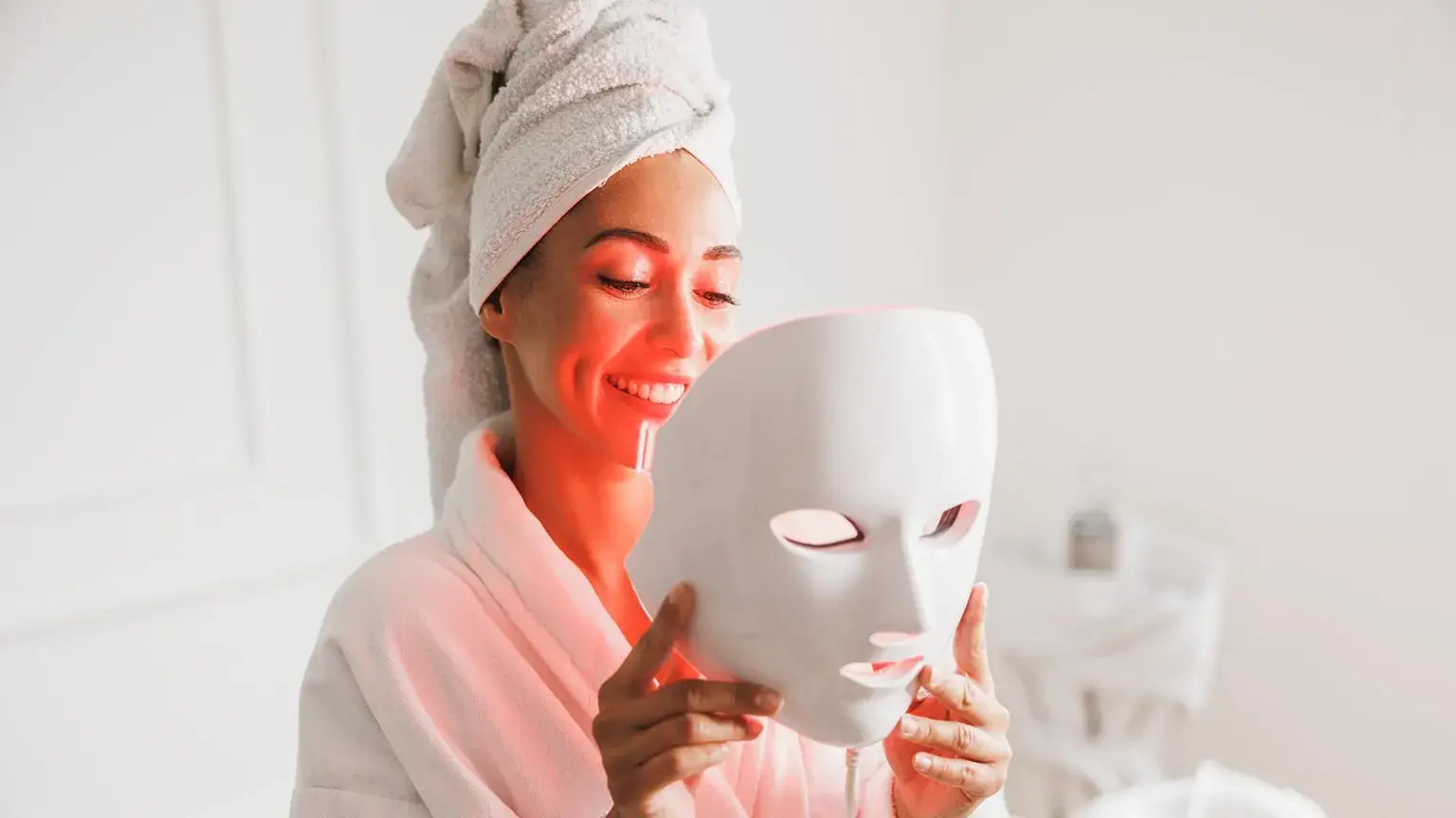 Woman in white robe holding LED face mask in bright bathroom setting