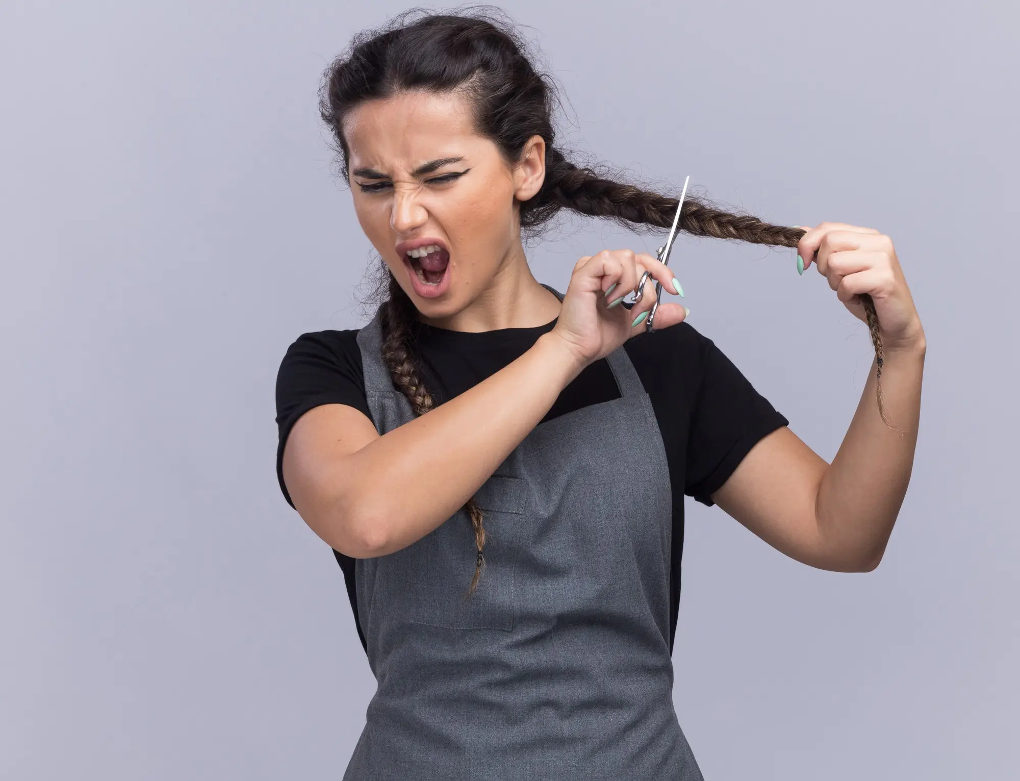 Person in gray apron cutting braided hair with scissors in front of plain background