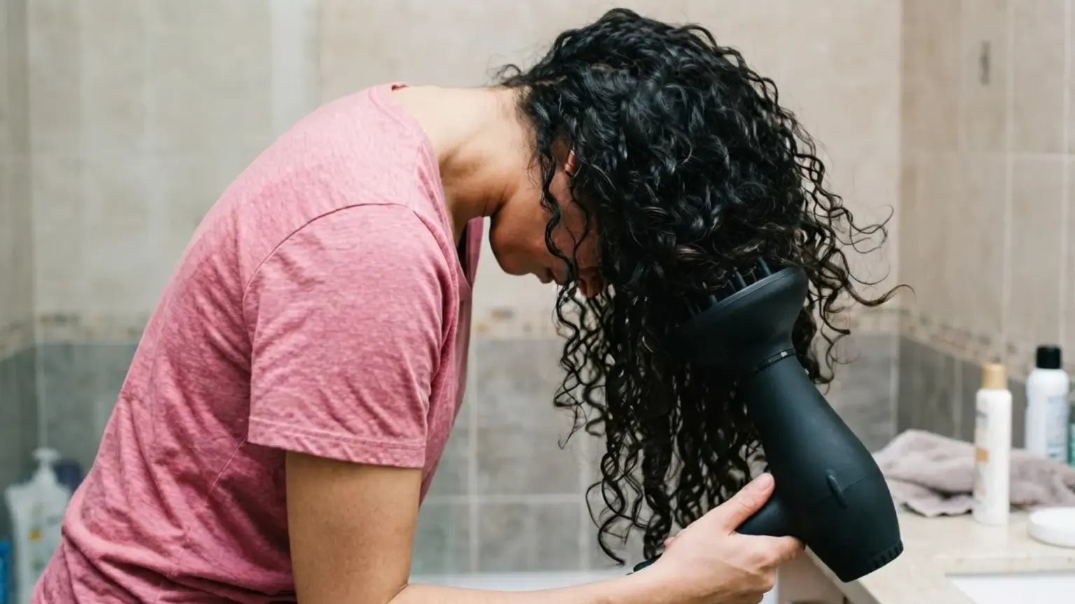A woman with curly hair uses a blow dryer to dry her hair, standing in a well-lit bathroom.