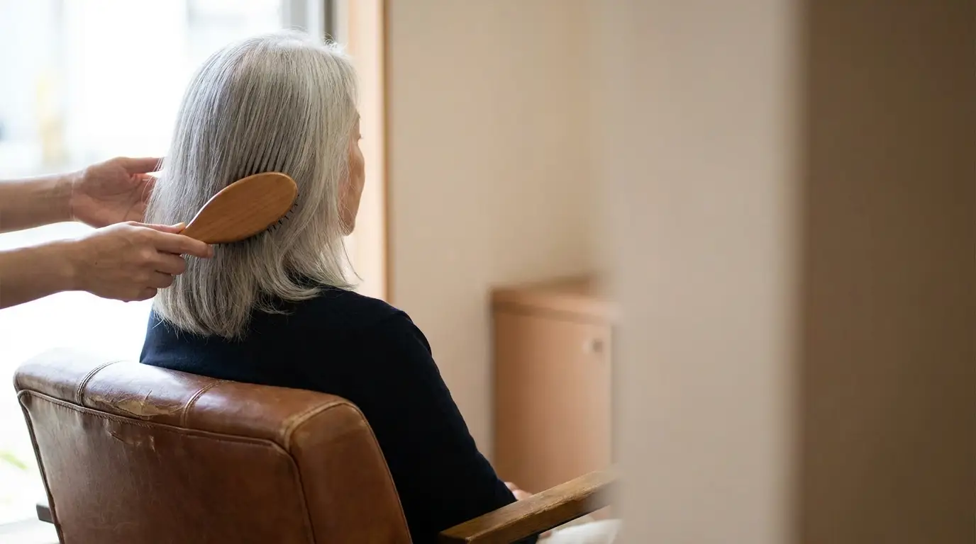 Gray-haired woman sitting in chair while someone brushes her hair indoors