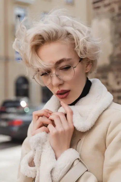 Woman with blonde hair and eyeglasses wearing beige coat outdoors in a city setting