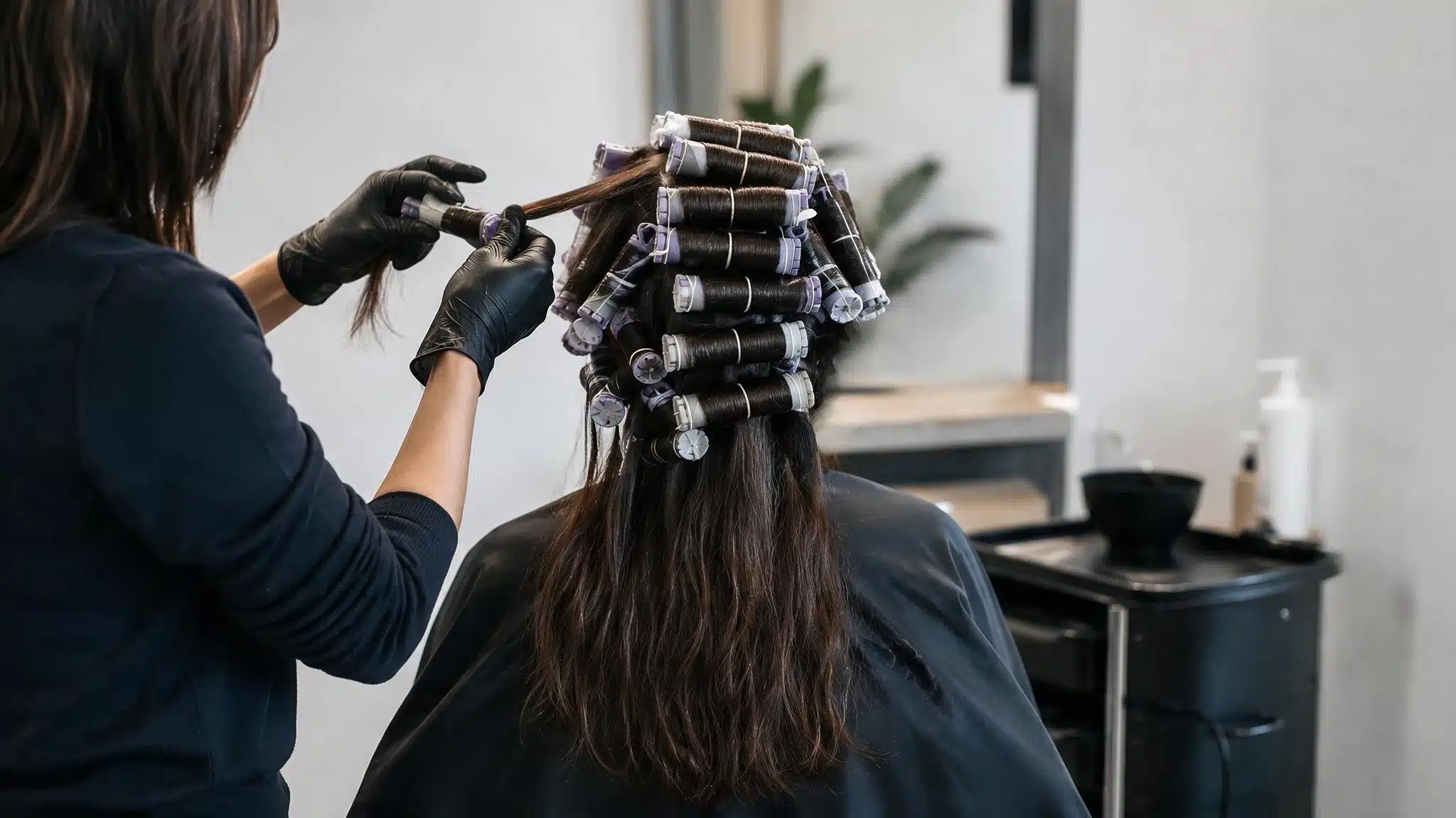 A woman sits in a salon chair, receiving a beach wave perm treatment from a stylist.