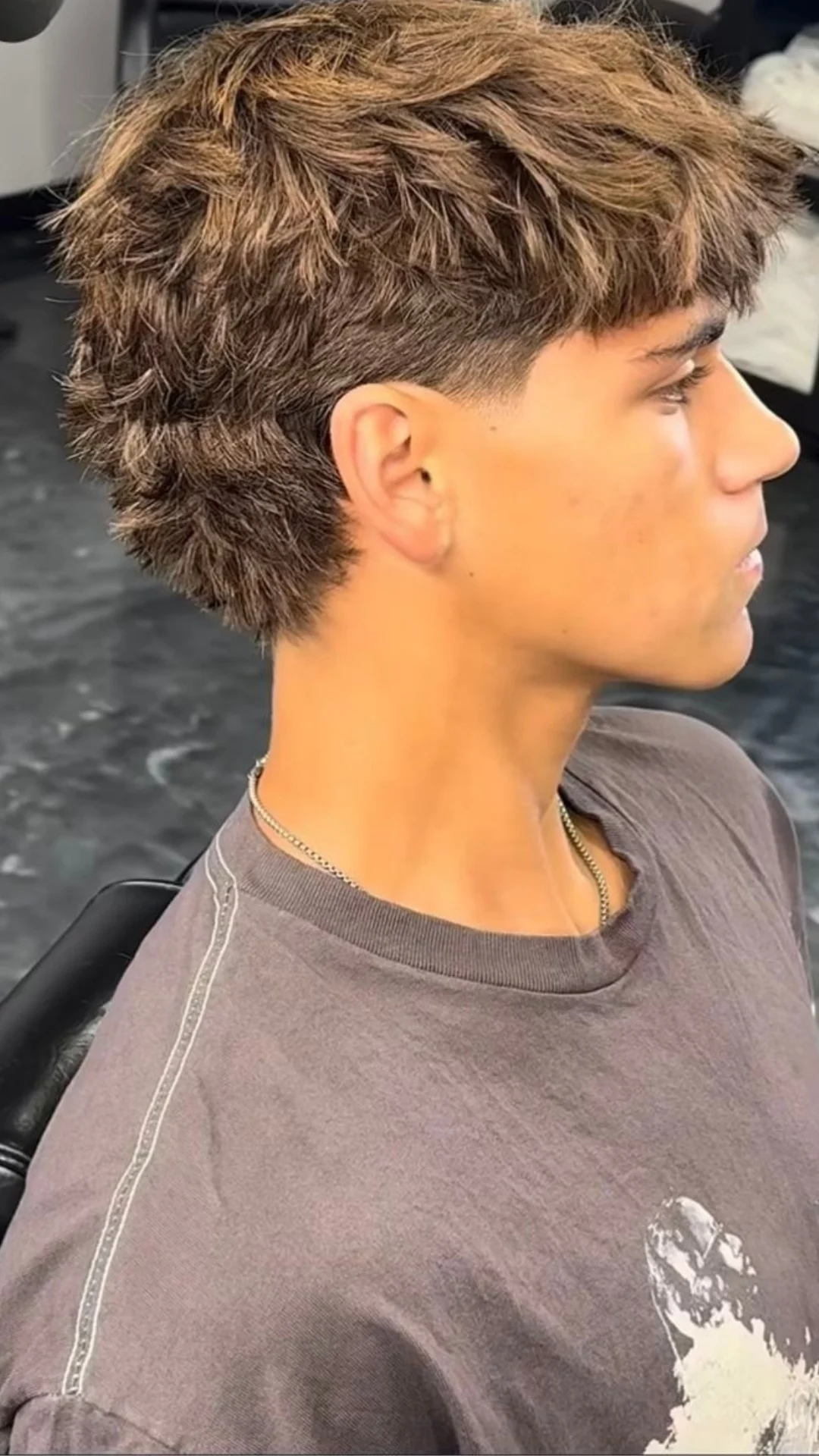 A young man with a short ice cream haircut sits in a salon chair, smiling at his new hairstyle.