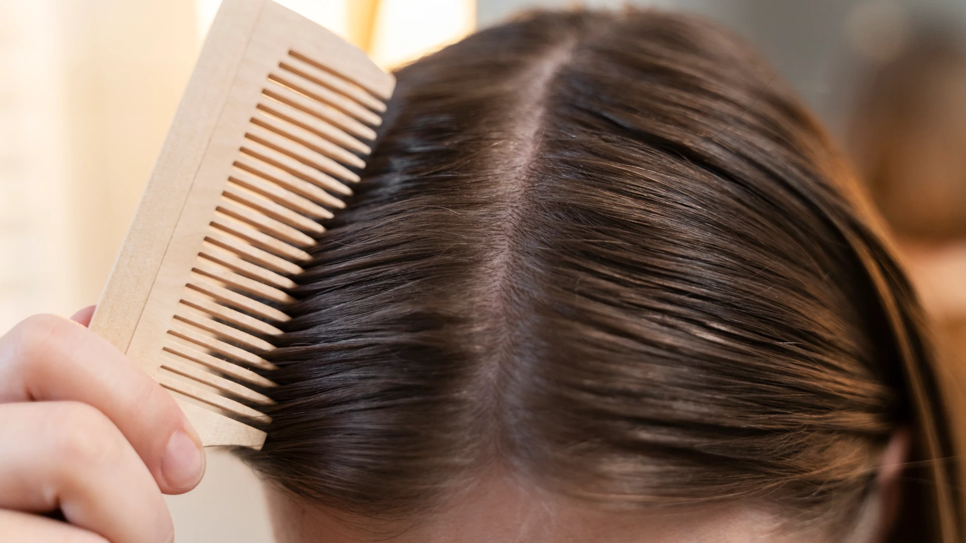 Close-up of a person using a wide-toothed comb to part and smooth dark brown hair, revealing the scalp.