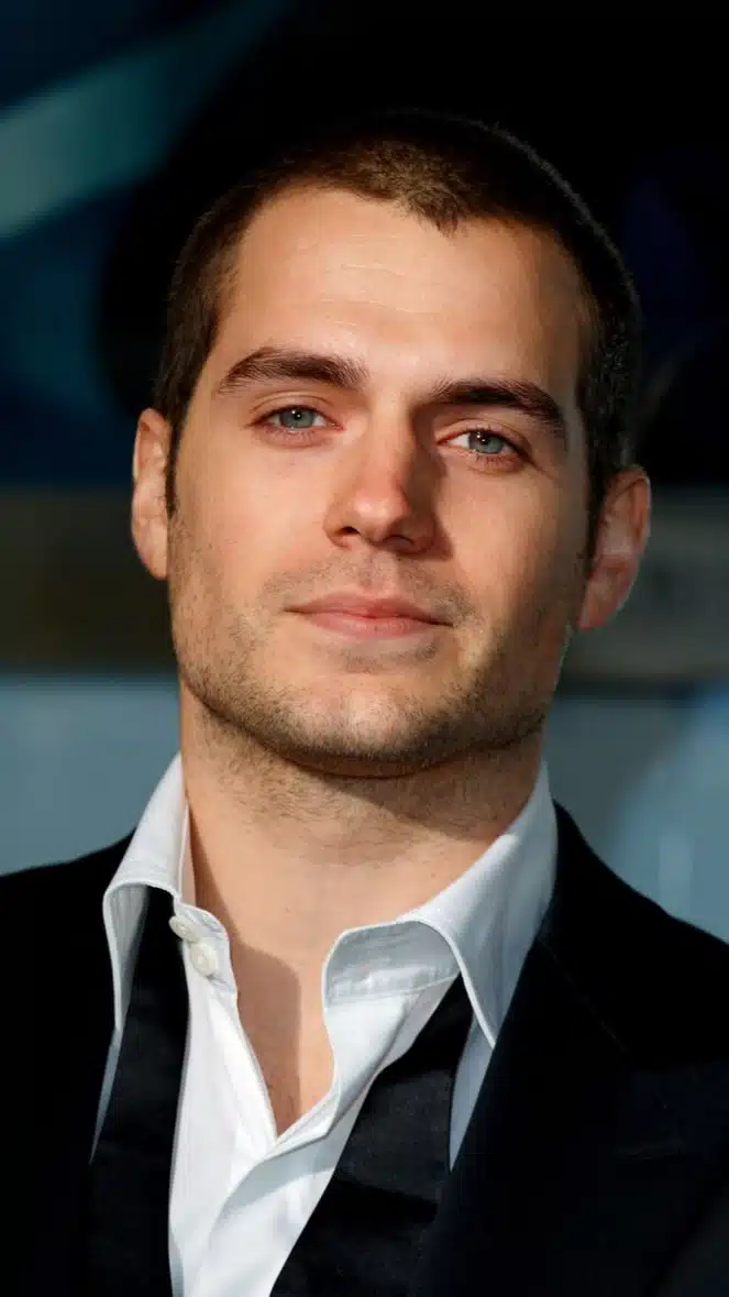 Henry Cavill in a suit and tie gazes confidently at the camera, exuding charm and sophistication.
