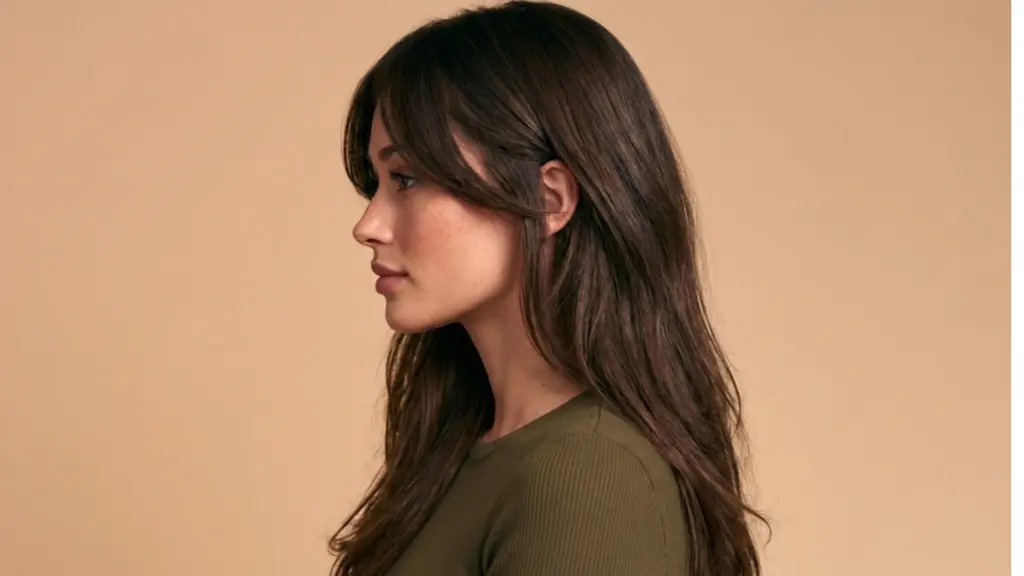 A woman with long hair with layered haircut dressed in a green top, posing confidently against a neutral background.