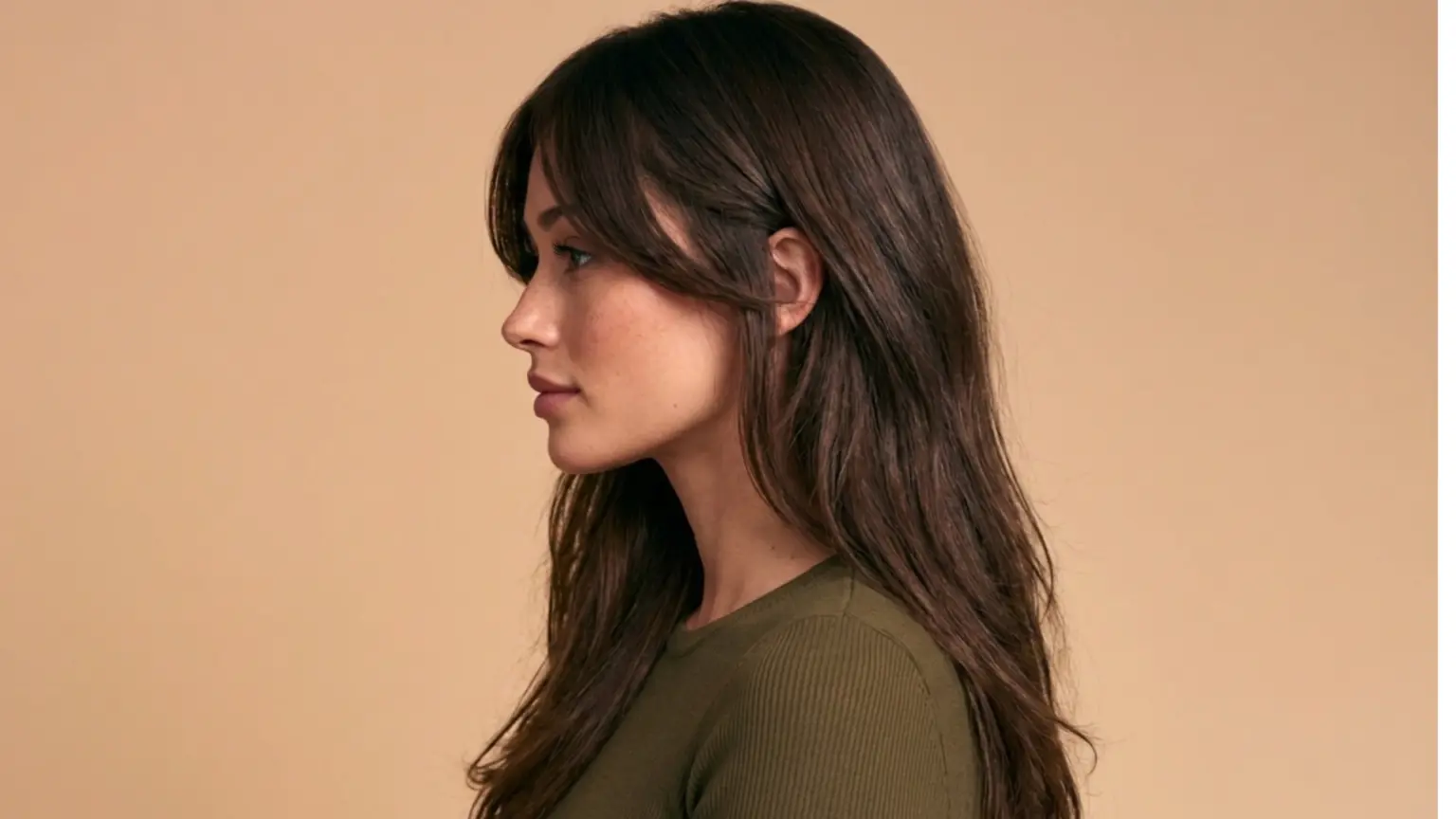 A woman with long hair with layered haircut dressed in a green top, posing confidently against a neutral background.