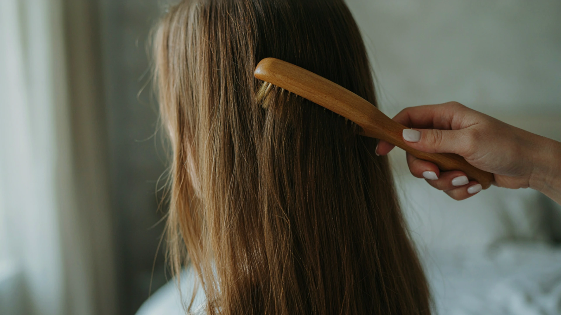Person brushing long hair with a wooden brush in a softly lit indoor setting.