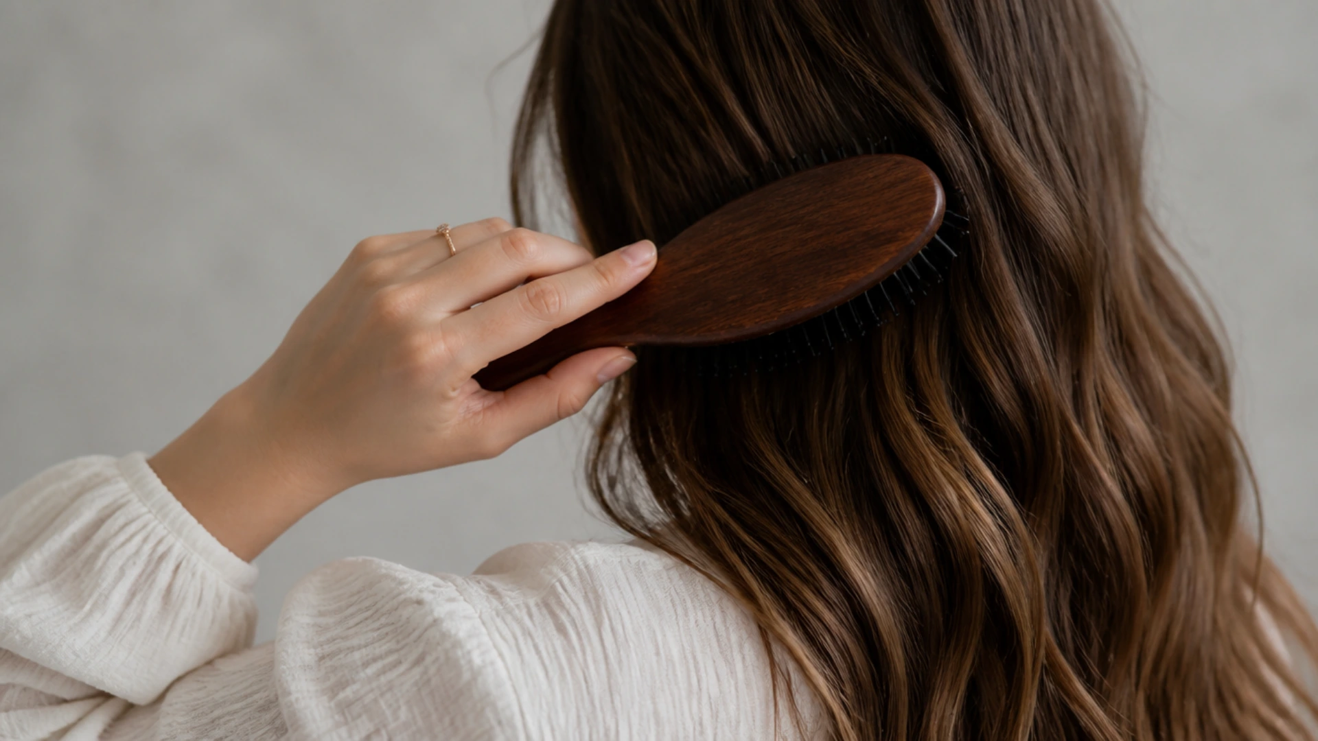 Person brushing long wavy hair with a wooden brush in a softly lit indoor environment.