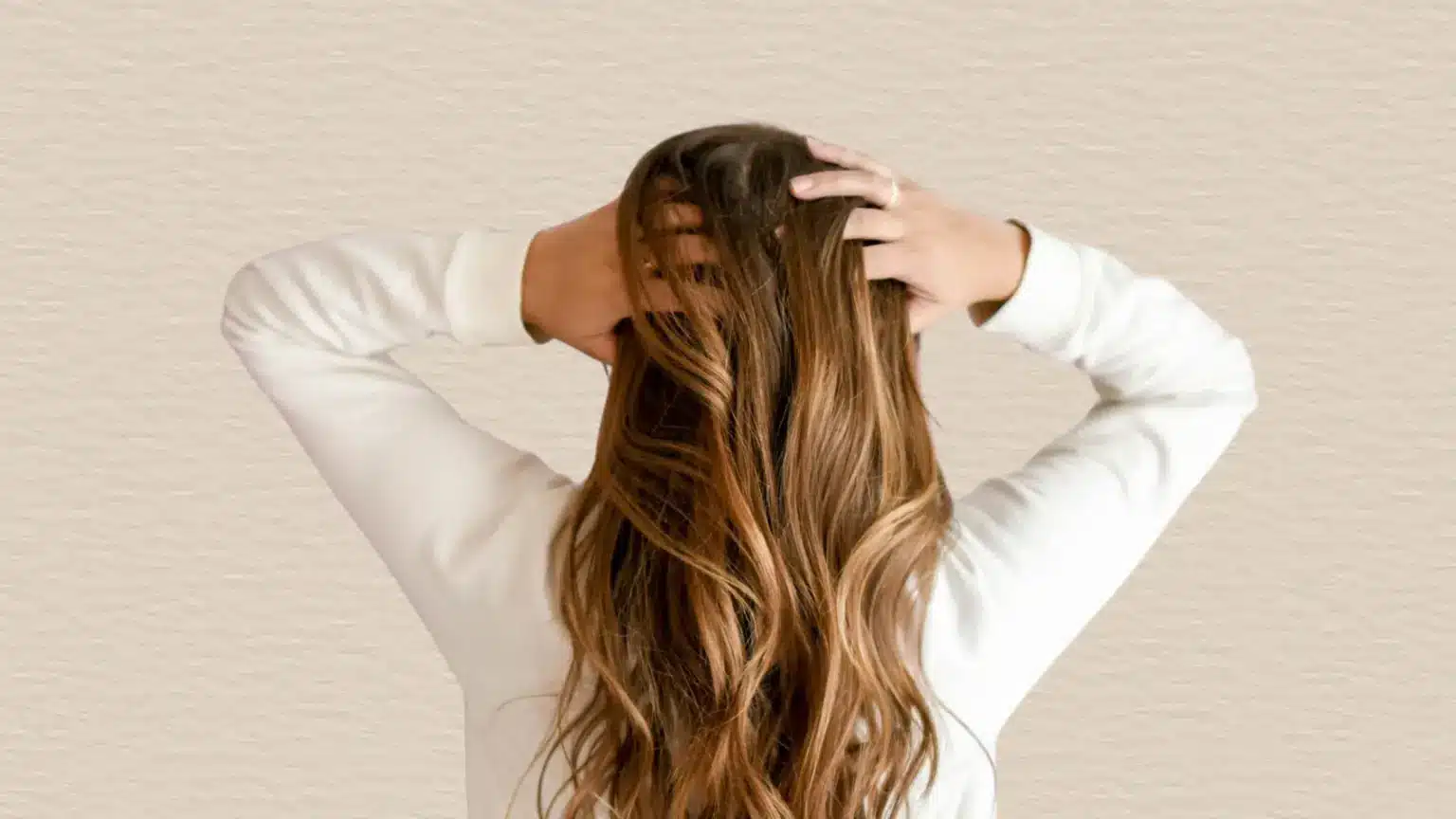 Back view of a woman with long brown wavy hair touching her scalp with both hands against a simple plain background